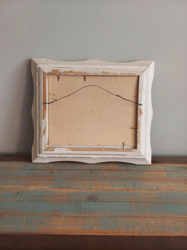 May include: White wooden picture frame with a scalloped edge. The frame is slightly distressed and has a wire hanger on the back.