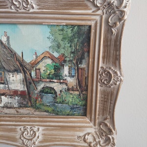 May include: A framed oil painting depicting a quaint village scene with houses, a bridge, and a stream. The painting is in a rustic, white-washed frame with ornate details.