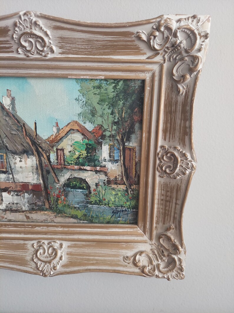 May include: A framed oil painting depicting a quaint village scene with houses, a bridge, and a stream. The painting is in a rustic, white-washed frame with ornate details.