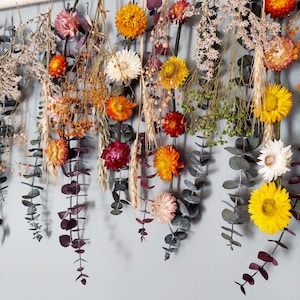 May include: A dried flower garland with a variety of colors, including yellow, orange, red, and white. The garland is made with dried flowers and eucalyptus leaves and is hanging from a wooden dowel.