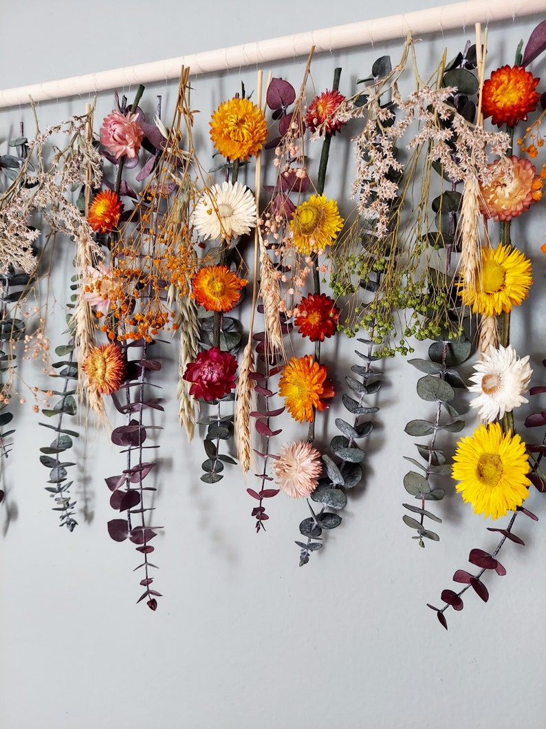 May include: A dried flower garland with a variety of colors, including yellow, orange, red, and white. The garland is made with dried flowers and eucalyptus leaves and is hanging from a wooden dowel.