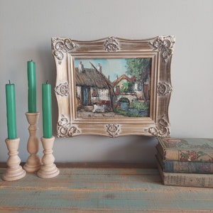 May include: A framed oil painting depicting a quaint village scene with houses, a bridge, and a river. The frame is ornate and has a distressed white finish.