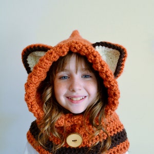 May include: A child wearing a crocheted fox hooded scarf with brown and orange stripes and a large brown button. The scarf has white and brown fox ears.