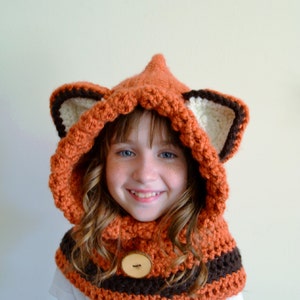 May include: A young person wearing a crocheted fox hat and scarf. The hat has brown and white ears and a brown button on the front. The scarf is orange and brown.