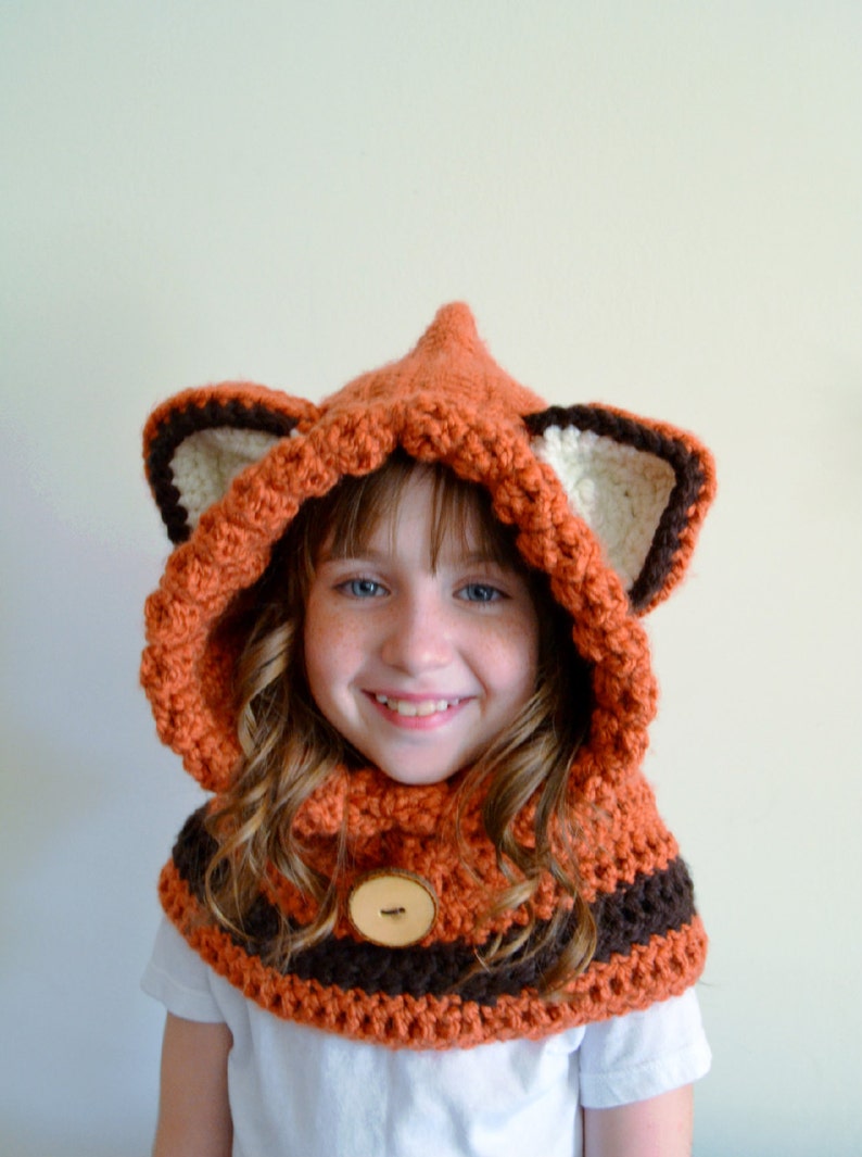 May include: A young person wearing a crocheted fox hat and scarf. The hat has brown and white ears and a brown button on the front. The scarf is orange and brown.