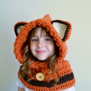 May include: A crocheted fox hat and scarf set in orange, brown, and white with a wooden button. The hat has a hood and large ears.