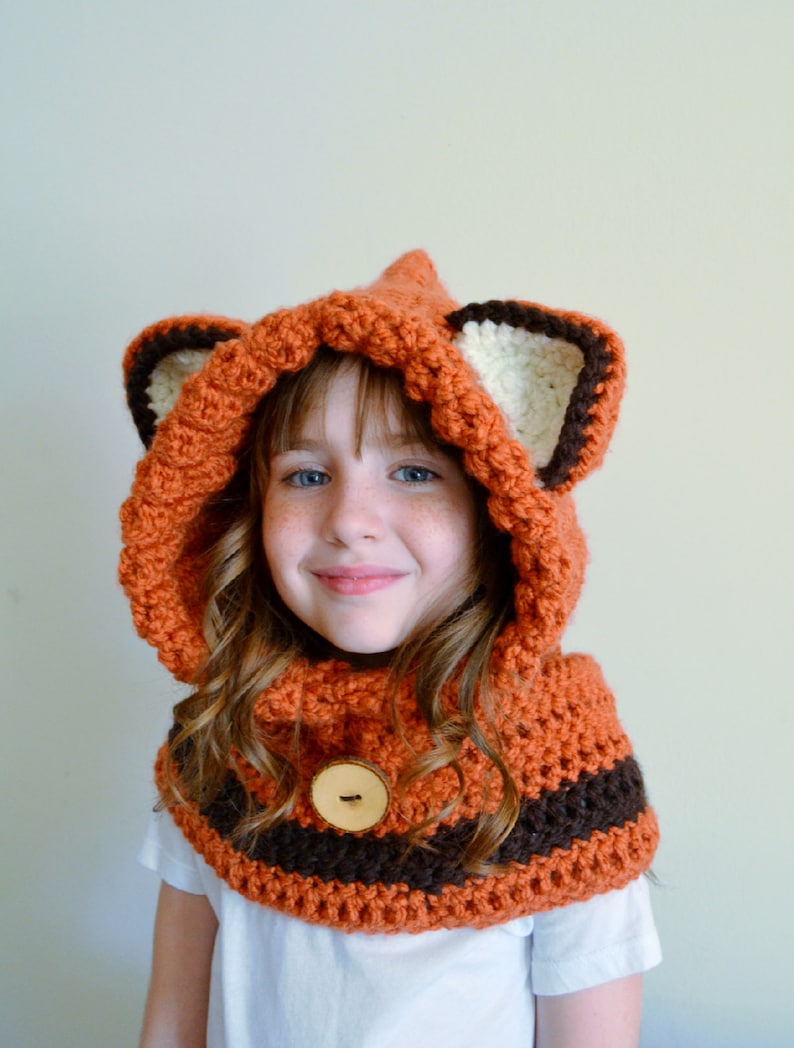 May include: A crocheted fox hat and scarf set in orange, brown, and white with a wooden button. The hat has a hood and large ears.
