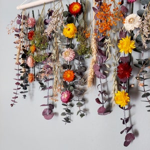 May include: A wall hanging made of dried flowers and greenery. The flowers are in shades of yellow, orange, red, and pink. The greenery is a mix of eucalyptus and other types of leaves.