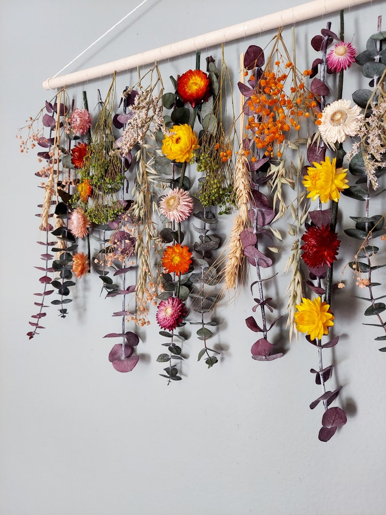 May include: A wall hanging made of dried flowers and greenery. The flowers are in shades of yellow, orange, red, and pink. The greenery is a mix of eucalyptus and other types of leaves.