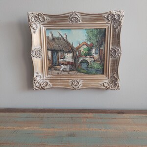 May include: A framed oil painting depicting a quaint village scene with houses, a bridge, and a river. The painting is in a gold-colored frame with ornate details.