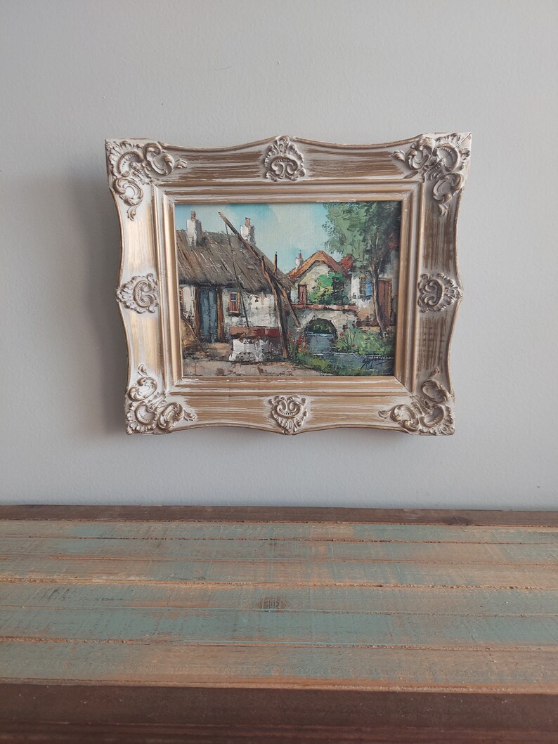 May include: A framed oil painting depicting a quaint village scene with houses, a bridge, and a river. The painting is in a gold-colored frame with ornate details.