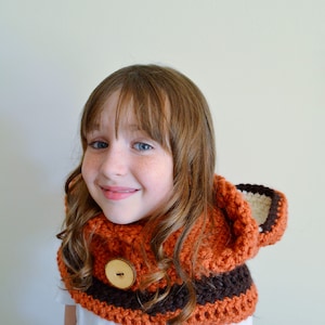 May include: A young person wearing a crocheted hooded cowl with a brown and orange color scheme. The cowl has a large wooden button on the front.