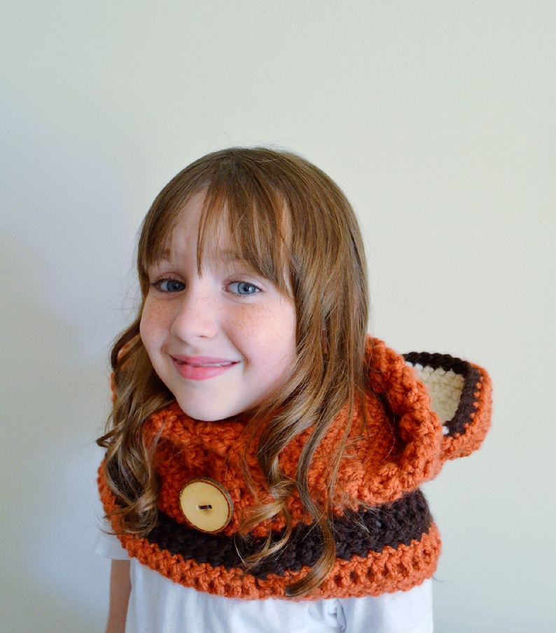 May include: A young person wearing a crocheted hooded cowl with a brown and orange color scheme. The cowl has a large wooden button on the front.