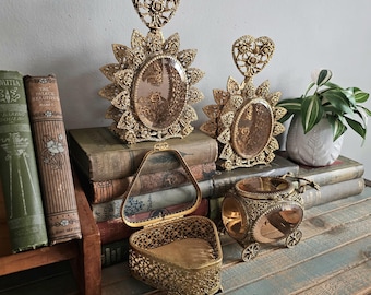 May include: Ornate gold-colored vanity items, including two perfume bottles with heart-shaped toppers and sunburst frames, a filigree heart-shaped trinket box, and a carriage-shaped container. The items are displayed on a vintage wooden surface.