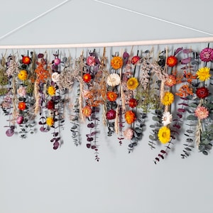 May include: A wall hanging made of dried flowers and eucalyptus leaves. The flowers are in shades of orange, yellow, red, and white. The leaves are a deep green. The hanging is attached to a wooden dowel.