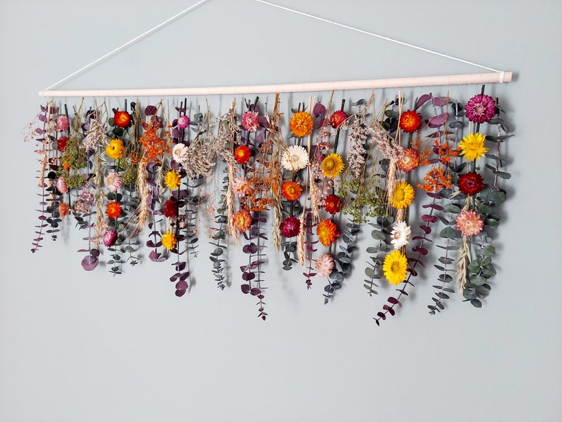 May include: A wall hanging made of dried flowers and eucalyptus leaves. The flowers are in shades of orange, yellow, red, and white. The leaves are a deep green. The hanging is attached to a wooden dowel.