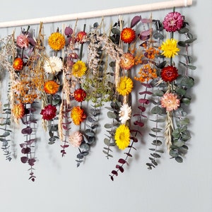 May include: A dried flower garland with a variety of colors, including orange, yellow, red, and purple. The garland is made with eucalyptus leaves and other dried flowers. It is hanging from a wooden dowel.