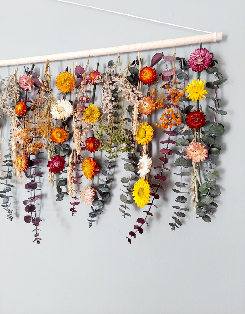 May include: A dried flower garland with a variety of colors, including orange, yellow, red, and purple. The garland is made with eucalyptus leaves and other dried flowers. It is hanging from a wooden dowel.