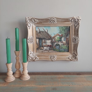 May include: A framed painting of a quaint village scene with houses, a bridge, and a river. The frame is ornate and has a gold finish. The painting is on a wooden surface with three candlesticks and candles.