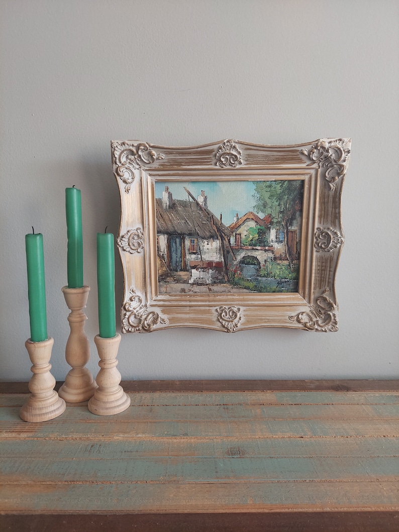 May include: A framed painting of a quaint village scene with houses, a bridge, and a river. The frame is ornate and has a gold finish. The painting is on a wooden surface with three candlesticks and candles.