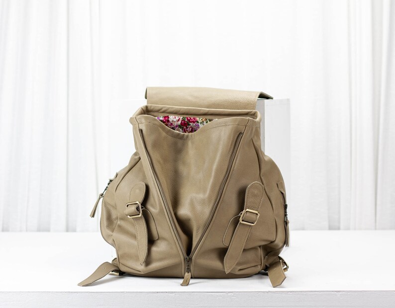 May include: A tan leather backpack with a floral lining. The backpack has two large zippered compartments and two smaller zippered pockets on the front.