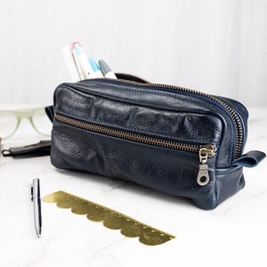 May include: A navy blue leather pencil case with a double zipper and a small handle. The case is open, revealing pens and markers. A gold-colored ruler and a silver pen are in the foreground. A pair of glasses and a notebook are in the background.