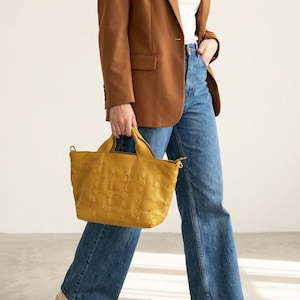 May include: A mustard yellow tote bag with a textured pattern is held by a person wearing a brown blazer and blue jeans. The bag has two handles and a shoulder strap. The person is walking in a well-lit space.