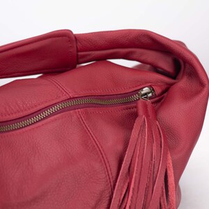K&ouml;nnte beinhalten: Eine rote Lederhandtasche mit Rei&szlig;verschluss und Quaste. Die Tasche hat einen Schulterriemen und ist aus weichem, strukturiertem Leder gefertigt. Der Rei&szlig;verschluss ist bronzefarben.