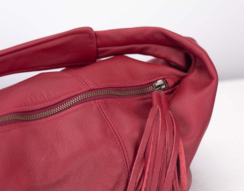 K&ouml;nnte beinhalten: Eine rote Lederhandtasche mit Rei&szlig;verschluss und Quaste. Die Tasche hat einen Schulterriemen und ist aus weichem, strukturiertem Leder gefertigt. Der Rei&szlig;verschluss ist bronzefarben.