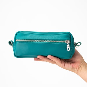 May include: Teal-colored leather cosmetic bag with a silver zipper closure. The rectangular bag has a small loop on each end and is held in a person's hand against a white background. The bag is approximately 8 inches long.