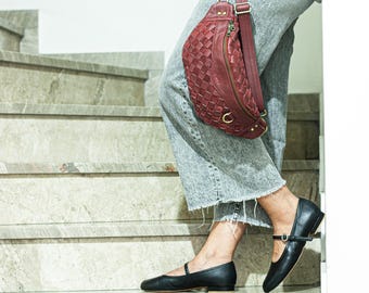 May include: A woman wearing grey jeans and black flats is carrying a red leather fanny pack with a woven design.
