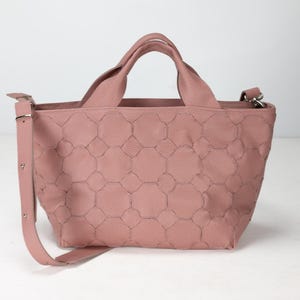 May include: A dusky pink tote bag with a geometric design. It features two short handles and a long, adjustable shoulder strap. The bag is made from a textured material and has a rectangular shape.