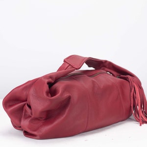 K&ouml;nnte beinhalten: Burgunderfarbene Lederhandtasche mit oberem Griff und Rei&szlig;verschluss. Die Tasche hat eine dekorative Fransenverzierung auf einer Seite. Das Leder wirkt weich und geschmeidig, mit leichtem Glanz. Die Tasche ist im l&auml;ssigen Stil gehalten.