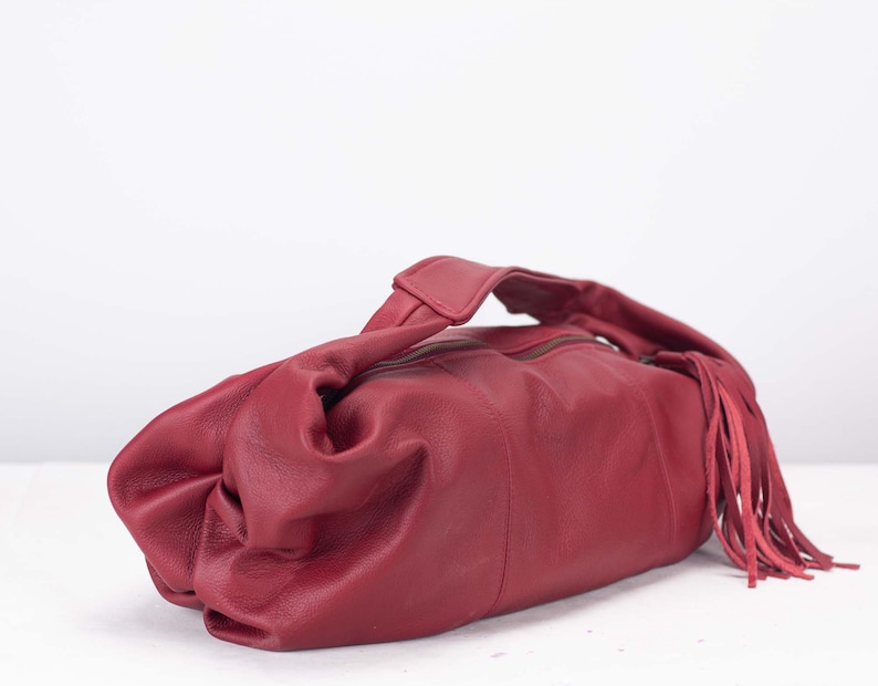 K&ouml;nnte beinhalten: Burgunderfarbene Lederhandtasche mit oberem Griff und Rei&szlig;verschluss. Die Tasche hat eine dekorative Fransenverzierung auf einer Seite. Das Leder wirkt weich und geschmeidig, mit leichtem Glanz. Die Tasche ist im l&auml;ssigen Stil gehalten.