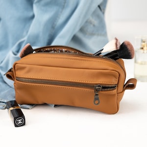 May include: A brown rectangular cosmetic bag with a zipper closure, containing makeup brushes. A black lipstick and a perfume bottle are also visible. The bag is made of a smooth material and has a zipper pull.