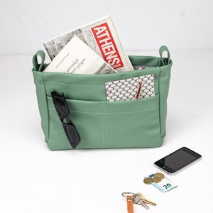 May include: A light green leather tote bag with multiple pockets, holding a newspaper with the word "ATHENS" visible, a book, and a patterned card. Sunglasses are in a pocket. A smartphone, keys, and money are next to the bag.