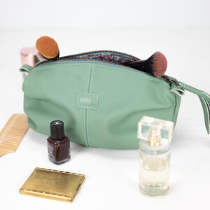 Op de afbeelding: Een lichtgroene leren toilettas met een ritssluiting en een kwast. De tas is open en bevat een make-upkwast, een flesje nagellak en een flesje parfum.