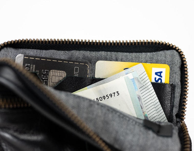 May include: A black leather zippered wallet with a gray fabric lining. The wallet is open and contains a black credit card, a yellow credit card, and a white card with the number 8095973.