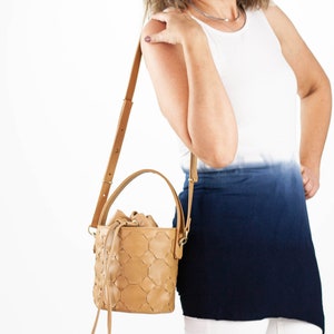 May include: A tan leather bucket bag with a woven pattern and a long strap. The bag is being worn by a person wearing a white and blue ombre tank top.