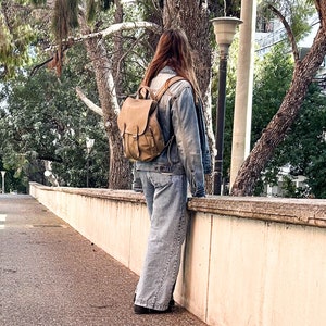 May include: A person wearing a denim jacket and blue jeans is standing in front of a stone wall. They are wearing a brown leather backpack with a buckle closure.
