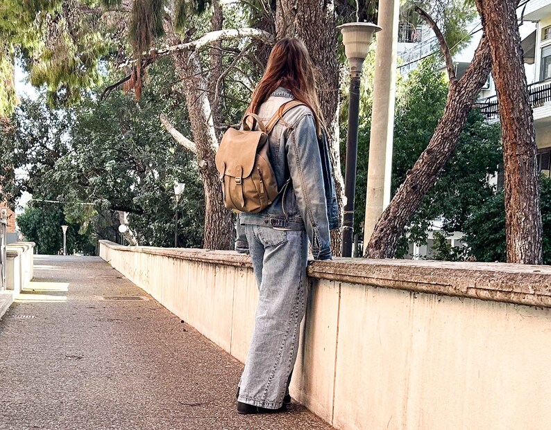 May include: A person wearing a denim jacket and blue jeans is standing in front of a stone wall. They are wearing a brown leather backpack with a buckle closure.