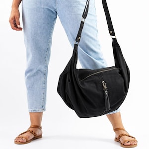 Op de afbeelding: Een zwarte schoudertas met ritssluiting en een kwastje. De tas is gemaakt van een zacht materiaal en heeft een verstelbare riem. De persoon op de foto draagt een lichtblauwe jeans en bruine sandalen.