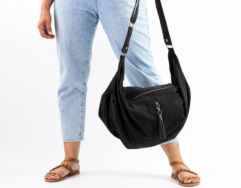 Op de afbeelding: Een zwarte schoudertas met ritssluiting en een kwastje. De tas is gemaakt van een zacht materiaal en heeft een verstelbare riem. De persoon op de foto draagt een lichtblauwe jeans en bruine sandalen.