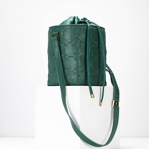 May include: A dark green leather bucket bag with a drawstring closure and a long shoulder strap. The bag has a geometric pattern embossed on the front.