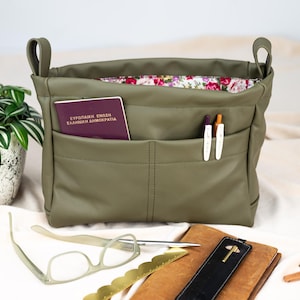 May include: Olive green tote bag with two handles and multiple front pockets. The interior lining features a floral pattern. A passport, pens, and other accessories are visible inside and around the bag. The bag is made of a smooth, durable material.