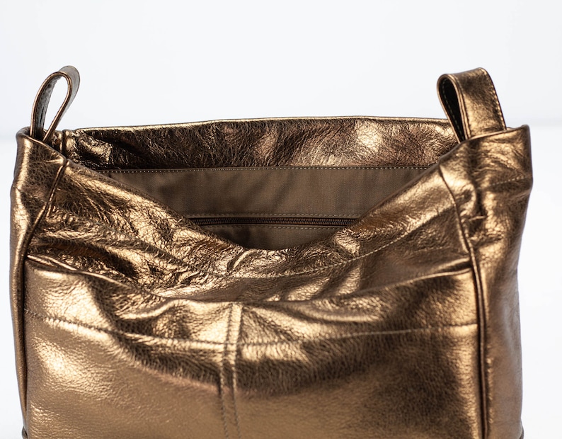 以下が含まれることがあります： 内側にジッパーポケットが付いたメタリックブロンズレザーのトートバッグ。バッグはゆったりとしたカジュアルなルックです。