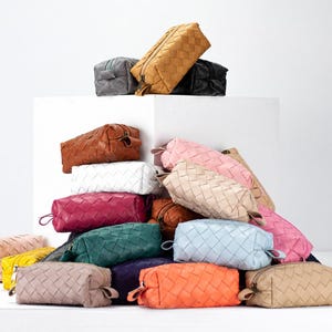 Pode incluir: Uma grande coleção de nécessaires retangulares com um padrão exterior entrançado. As bolsas estão disponíveis numa variedade de cores sólidas, incluindo amarelo mostarda, branco, magenta, azul-petróleo, azul-claro, laranja, bege, rosa e preto.