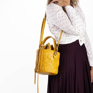 May include: A yellow bucket bag with a textured design and a long strap. The bag is worn over the shoulder of a person wearing a white polka dot shirt and a burgundy skirt.