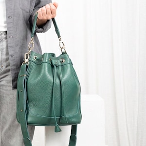 May include: A green leather bucket bag with a drawstring closure and a shoulder strap. The bag has gold-coloured hardware and a textured surface. The bag is held by a person.