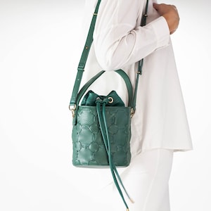 May include: A green quilted leather bucket bag with a gold drawstring closure and a long adjustable strap. The bag is worn over the shoulder of a person wearing a white shirt.